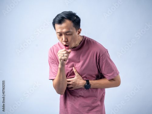 A man in a pink shirt coughs into his fist while holding his chest, indicating discomfort. He wears a black wristwatch. The background is a plain, light blue backdrop