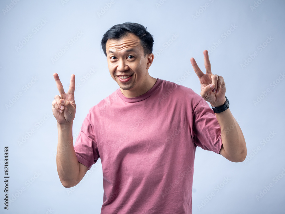 An Asian man in a pink shirt smiles and flashes a peace sign with both hands, exuding a cheerful and relaxed demeanor. The plain light blue background highlights his positive and friendly expression. An Asian man in a pink shirt smiles and flashes a peace sign with both hands, exuding a cheerful and relaxed demeanor. The plain light blue background highlights his positive and friendly expression.