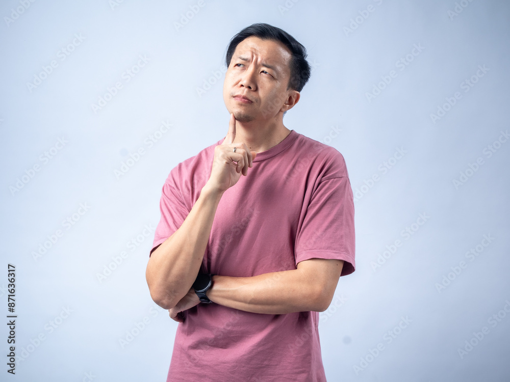 A man in a pink shirt looks thoughtfully upwards with his hand on his chin, reflecting a pensive and contemplative mood. He stands against a plain light blue background A man in a pink shirt looks thoughtfully upwards with his hand on his chin, reflecting a pensive and contemplative mood. He stands against a plain light blue background