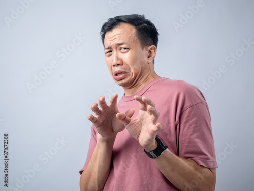 A man in a pink shirt makes a disgusted and fearful expression with his hands raised in a defensive gesture. The plain light blue background emphasizes his reaction photo