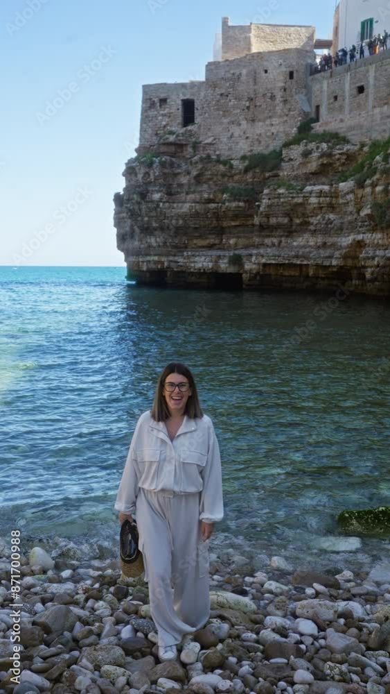 A beautiful young hispanic woman stands by the rocky beach in polignano a mare, puglia, italy, with clear blue waters and ancient stone buildings in the background. A beautiful young hispanic woman stands by the rocky beach in polignano a mare, puglia, italy, with clear blue waters and ancient stone buildings in the background.