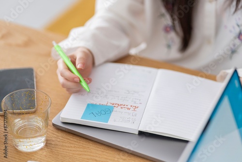 Close-up of a person writing notes in a notebook at a desk with a green pen A glass and a smartphone are also on the desk photo