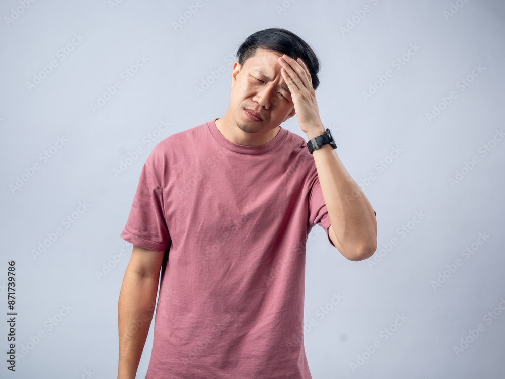 A man in a pink shirt holds his head with a pained expression, indicating a headache or discomfort. The plain light blue background emphasizes his distress and highlights his posture and expression. A man in a pink shirt holds his head with a pained expression, indicating a headache or discomfort. The plain light blue background emphasizes his distress and highlights his posture and expression.