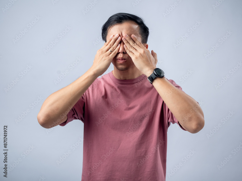 A man in a pink shirt covers his eyes with his hands while screaming, showing an expression of surprise or shock. The plain light blue background emphasizes his dramatic and emotional reaction. A man in a pink shirt covers his eyes with his hands while screaming, showing an expression of surprise or shock. The plain light blue background emphasizes his dramatic and emotional reaction.