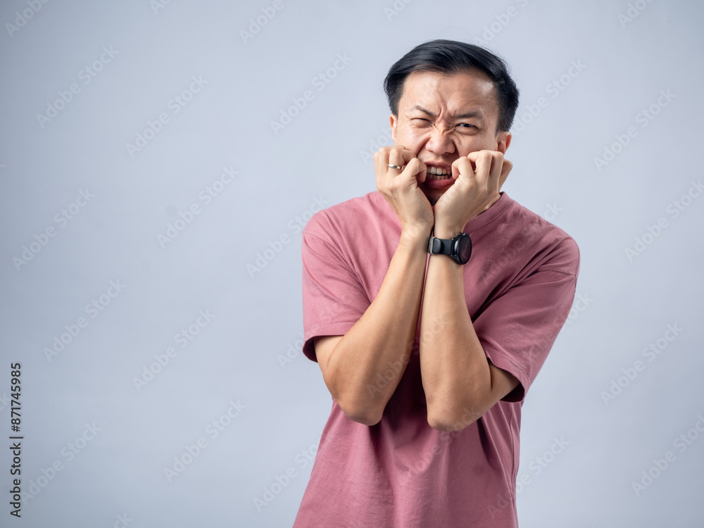 Asian man in a pink t-shirt stands against a plain background, displaying a nervous and anxious expression. He has his hands near his mouth, and his posture and facial expression Asian man in a pink t-shirt stands against a plain background, displaying a nervous and anxious expression. He has his hands near his mouth, and his posture and facial expression