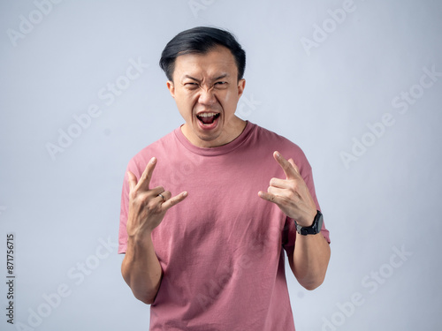 An Asian man in a pink t-shirt makes rock and roll hand gestures, shouting with enthusiasm. The light grey studio background highlights his energetic and rebellious expression