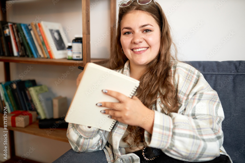 Indoor portrait of pretty smiling plus size girl with glasses on forehead writing ideas, plans for life or memories in her diary sitting on couch at home with pen and copybook in hand Indoor portrait of pretty smiling plus size girl with glasses on forehead writing ideas, plans for life or memories in her diary sitting on couch at home with pen and copybook in hand