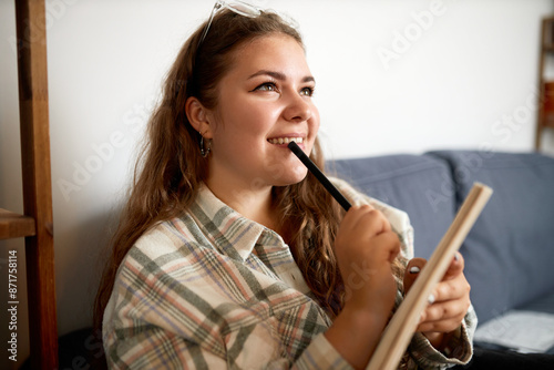 Side view of romantic dreamy thoughtful young lady writer beginner with glasses on head inventing new plot for her story or novel, sitting on sofa touching lips with pan and holding copybook