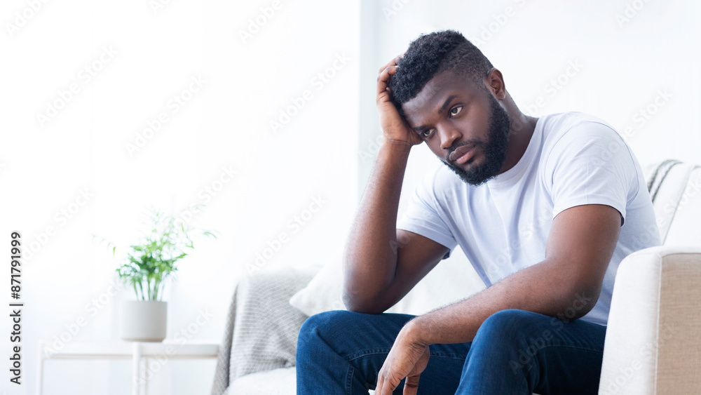 A young black man, with a beard, sits on a white couch in a modern apartment. He is wearing a white t-shirt and blue jeans. He has his head in his hand and a pensive expression on his face. A young black man, with a beard, sits on a white couch in a modern apartment. He is wearing a white t-shirt and blue jeans. He has his head in his hand and a pensive expression on his face.