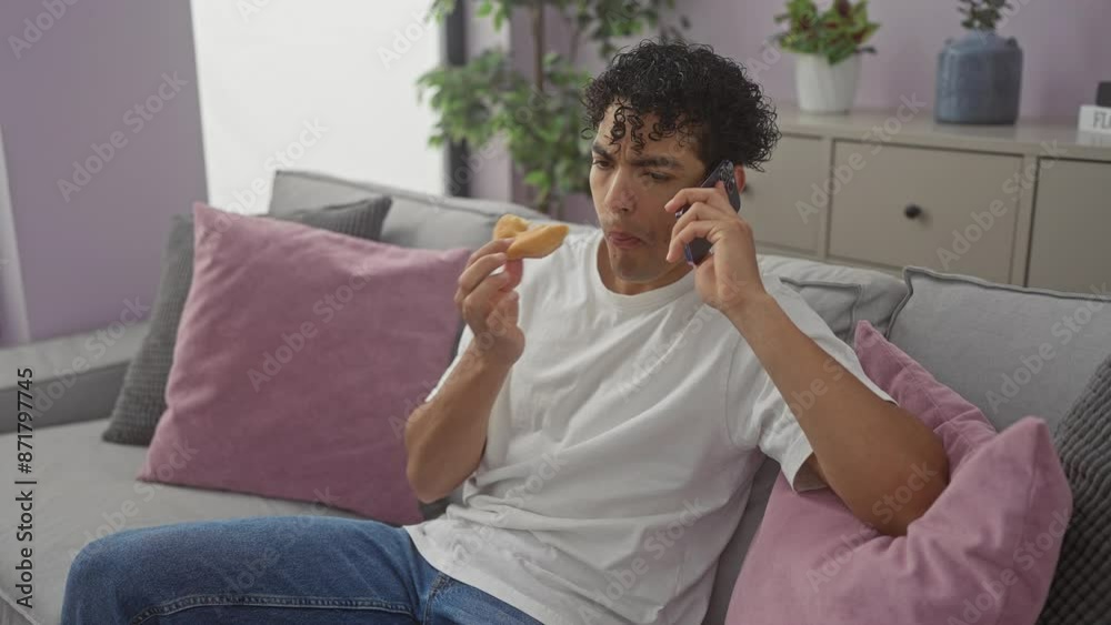 A young hispanic man in casual wear multitasks at home, talking on the phone while eating a pastry on a cozy couch. A young hispanic man in casual wear multitasks at home, talking on the phone while eating a pastry on a cozy couch.