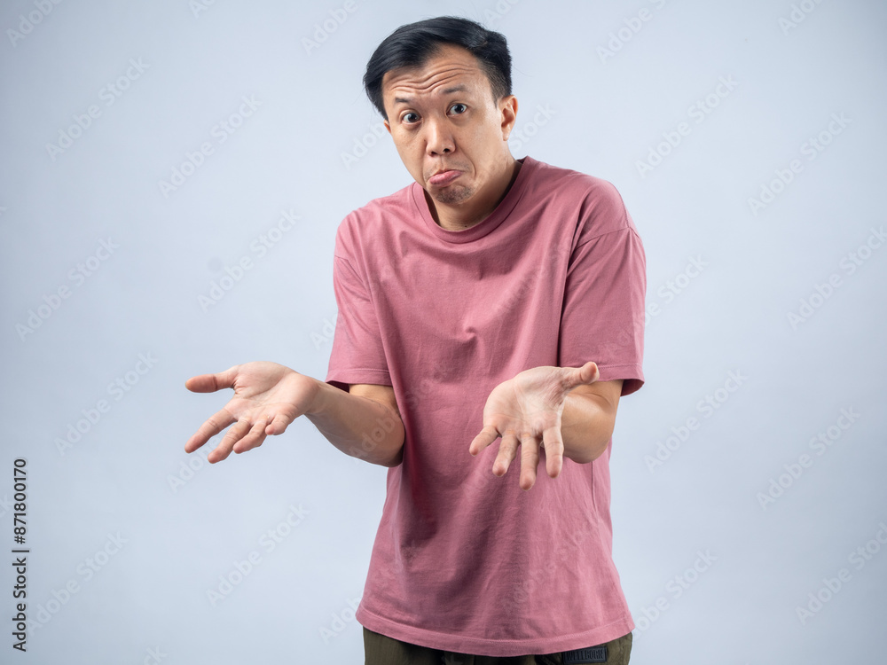 Naklejka premium An Asian man in a pink shirt shrugs with a confused and uncertain expression, his arms outstretched and palms up. The plain light blue background emphasizes his ambiguous