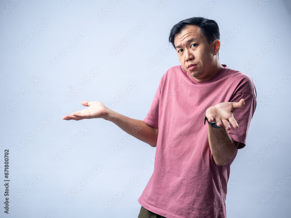 Naklejka premium An Asian man in a pink shirt shrugs with a confused and uncertain expression, his arms outstretched and palms up. The plain light blue background emphasizes his ambiguous