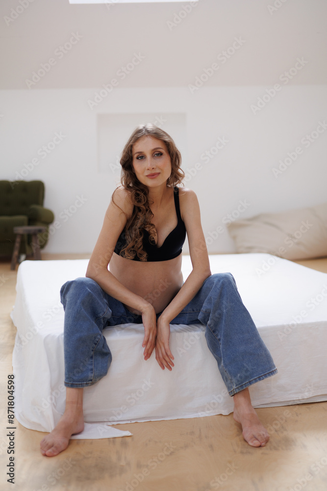 Beautiful pregnant woman in black top and jeans barefoot, sitting. Big pregnant naked belly. Pretty woman waiting child. Sofa, small window, white walls Beautiful pregnant woman in black top and jeans barefoot, sitting. Big pregnant naked belly. Pretty woman waiting child. Sofa, small window, white walls