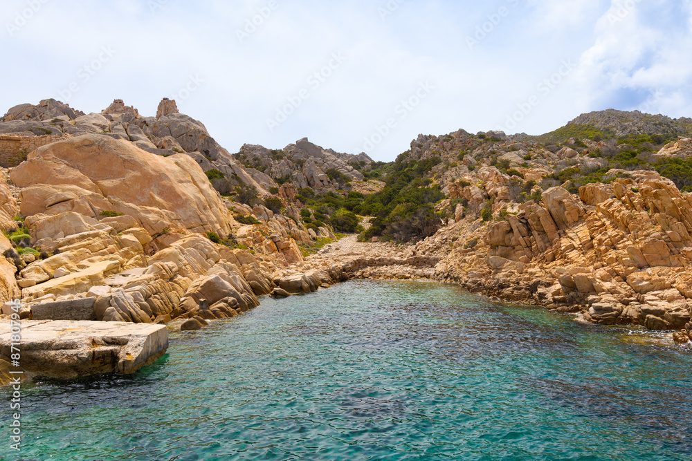 Fototapeta premium Cala Candeo en la isla de Caprera, parte del archipiélago de la Magdalena, Cerdeña, Italia. Una pequeña cala rodeada de formaciones rocosas de tonos anaranjados y vegetación mediterránea.