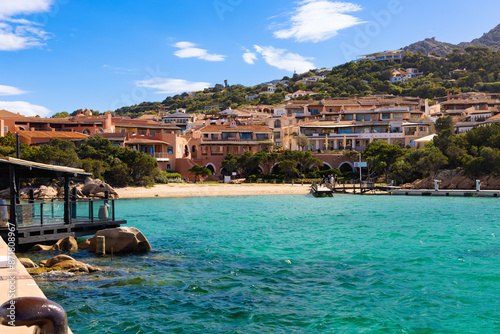 Vista de Porto Cervo en Cerdeña, Italia, en un día soleado. Las aguas turquesas del mar rodean un muelle y una playa de arena, con villas de color terracota construidas en la ladera. 