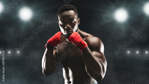 A boxer in red hand wraps stands ready in a dark stadium, illuminated by bright lights. photo