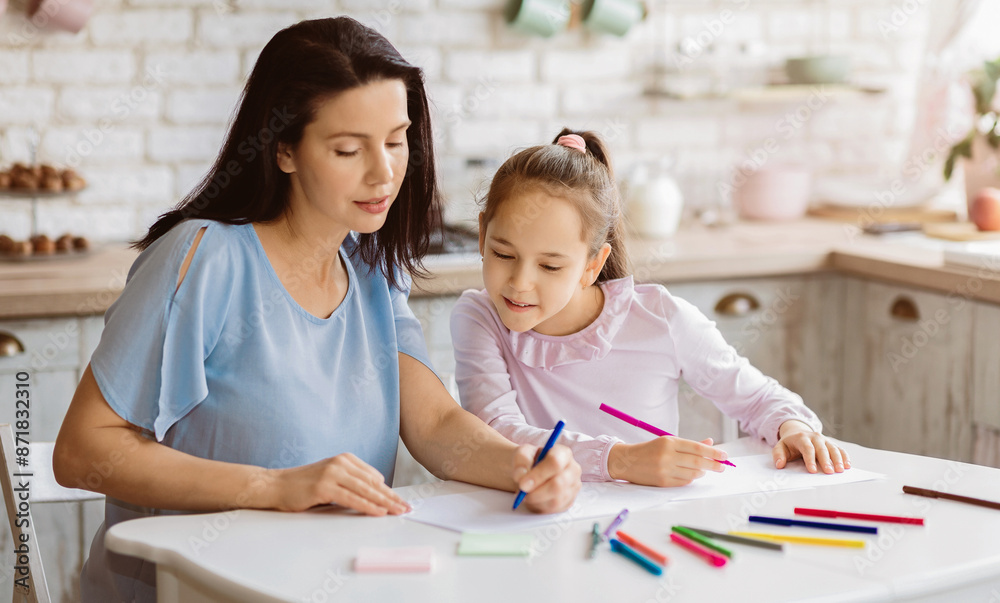 A mother and her young daughter are sitting at a kitchen table, both focused on drawing with colored pencils. They are both smiling, suggesting a happy and enjoyable experience. A mother and her young daughter are sitting at a kitchen table, both focused on drawing with colored pencils. They are both smiling, suggesting a happy and enjoyable experience.