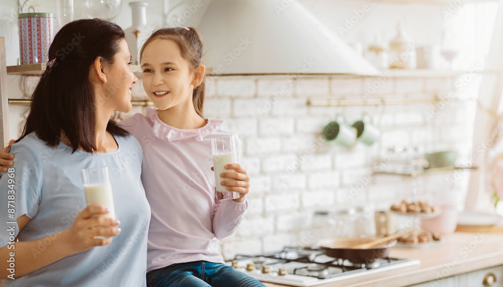 A mother and her young daughter stand side by side in a kitchen, both holding glasses of milk. They are smiling at each other and appear to be enjoying their time together
