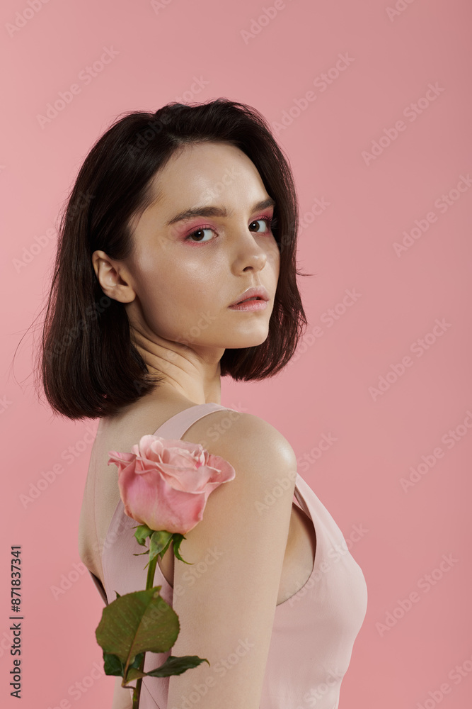 A woman with short dark hair poses with a pink rose against a pink backdrop. A woman with short dark hair poses with a pink rose against a pink backdrop.