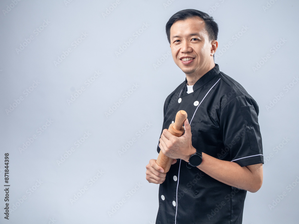 Fototapeta premium A smiling Asian male chef in a black chef's uniform, holding a wooden rolling pin, exudes friendliness and readiness for baking tasks against a light blue background
