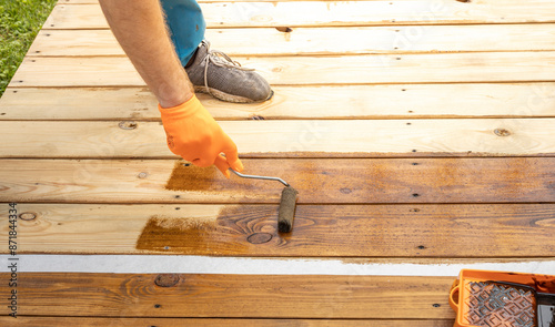 Wallpaper Mural A persons gloved hand uses a roller to apply stain to wooden deck boards. The stain is already applied to half the deck, showing the difference in color. Torontodigital.ca