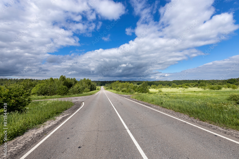 A straight, paved road winds through a lush green field, under a partly cloudy blue sky.. A straight, paved road winds through a lush green field, under a partly cloudy blue sky..
