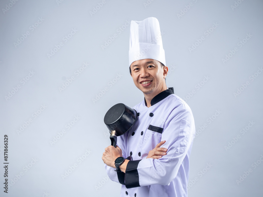 A confident Asian male chef in a white chef's uniform and hat, holding a pan across his chest, smiles warmly against a light blue background. The setting is culinary and professional. A confident Asian male chef in a white chef's uniform and hat, holding a pan across his chest, smiles warmly against a light blue background. The setting is culinary and professional.