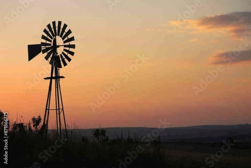 An old windmill silhouetted against a sunset sky, with a softly blurred background of fields and distant hills. 