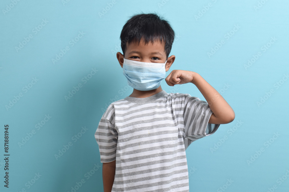 Asian Boy wearing health mask isolated on blue background. Confident kid shows his biceps arm muscle. Medical and health concept Asian Boy wearing health mask isolated on blue background. Confident kid shows his biceps arm muscle. Medical and health concept