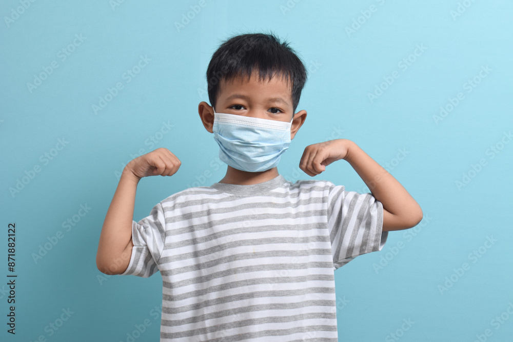 Asian Boy wearing health mask isolated on blue background. Confident kid shows his biceps arm muscle. Medical and health concept Asian Boy wearing health mask isolated on blue background. Confident kid shows his biceps arm muscle. Medical and health concept