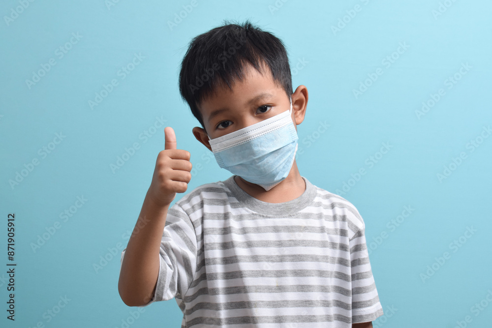 Asian Boy wearing a health mask while giving a thumbs up isolated on a blue background. Medical and health concept. Human population viruses, infections, flu disease Asian Boy wearing a health mask while giving a thumbs up isolated on a blue background. Medical and health concept. Human population viruses, infections, flu disease