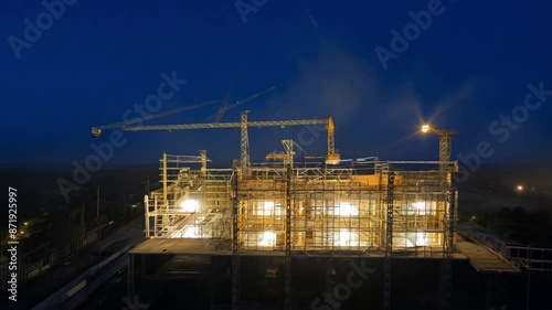 Aerial view of the time-lapse At a construction site plant surrounded by industrial architecture at night, steel structures and cranes are on display against a deep blue sky.