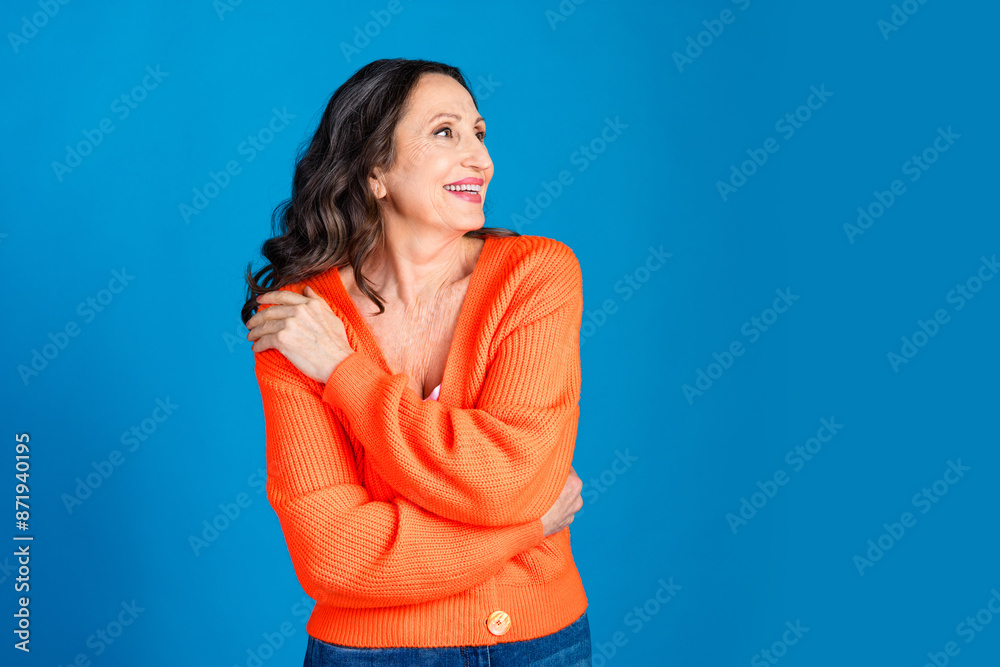 Photo portrait of attractive senior woman embrace self look empty space dressed stylish orange clothes isolated on blue color background Photo portrait of attractive senior woman embrace self look empty space dressed stylish orange clothes isolated on blue color background