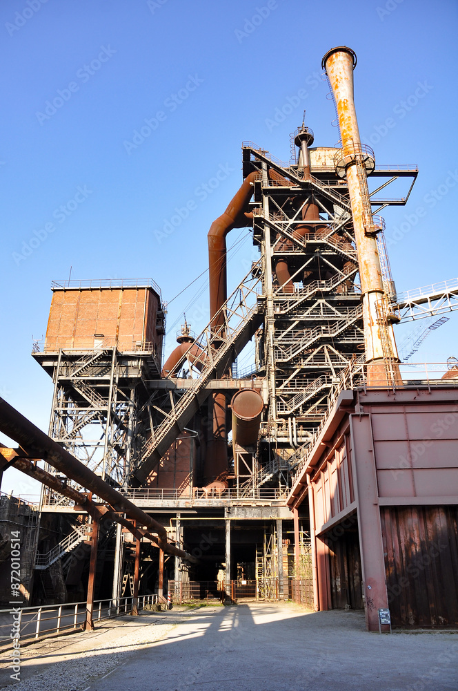 The photograph depicts an old metallurgical plant in Duisburg. Rusty metal structures and pipes dominate the foreground, highlighting the industrial heritage of heavy steel production. The factory's c The photograph depicts an old metallurgical plant in Duisburg. Rusty metal structures and pipes dominate the foreground, highlighting the industrial heritage of heavy steel production. The factory's c