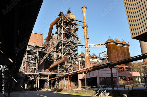 The photograph depicts an old metallurgical plant in Duisburg. Rusty metal structures and pipes dominate the foreground, highlighting the industrial heritage of heavy steel production. The factory's c