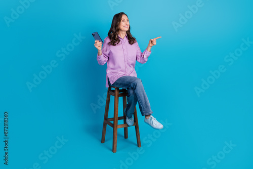 Full length photo of cheerful pretty lady dressed puple blouse sitting stool typing modern device pointing empty space isolated blue color background photo