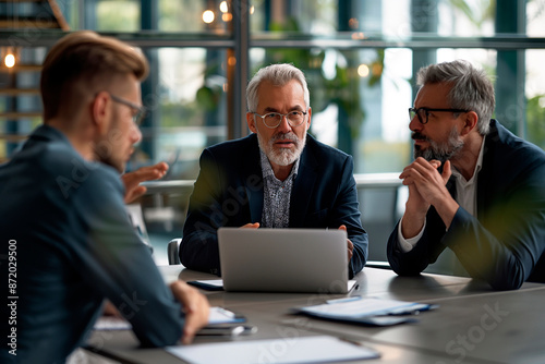 Middle aged businessman leading a meeting with two colleagues in a modern office in a discussion with laptop and notes on the table