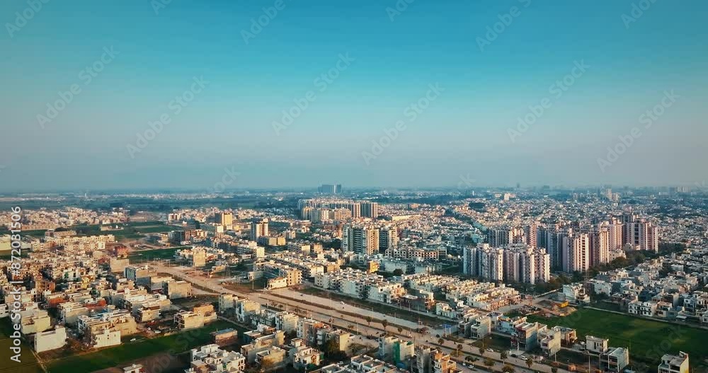 Hovering above urban structures, a drone moves towards the horizon, capturing Mohali city at sunset with a blue sky and yellowish buildings creating an amazing hue. Hovering above urban structures, a drone moves towards the horizon, capturing Mohali city at sunset with a blue sky and yellowish buildings creating an amazing hue.