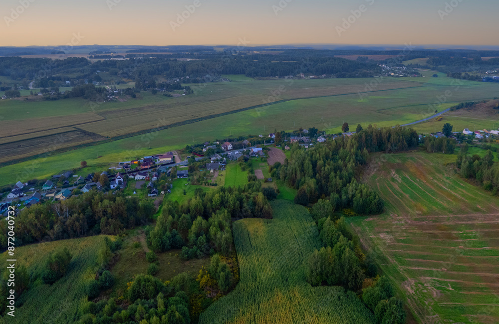 Obraz premium Suburban houses in village, aerial view. Rural landscape at evening sunset. Village wooden house in rural. Roofs of village houses in countryside, drone view. Suburb house at farm field, aerial view.