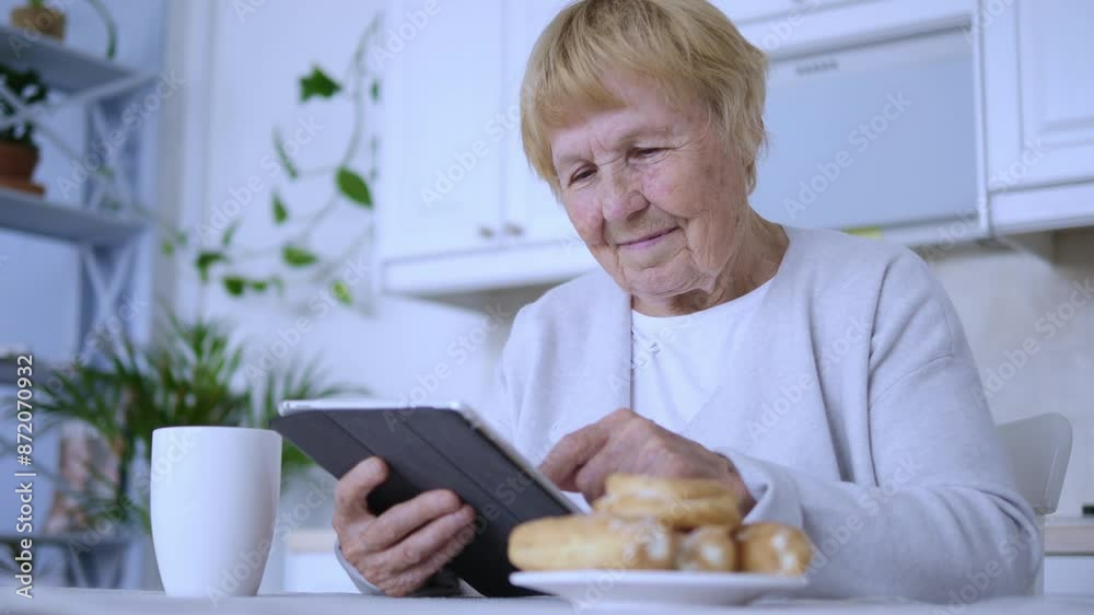 Elderly grandmother sits at kitchen table using tablet with cup of coffee and pastries nearby. Senior grandmother pensioner comfortably seated at kitchen table browsing on tablet with cup of coffee. Elderly grandmother sits at kitchen table using tablet with cup of coffee and pastries nearby. Senior grandmother pensioner comfortably seated at kitchen table browsing on tablet with cup of coffee.