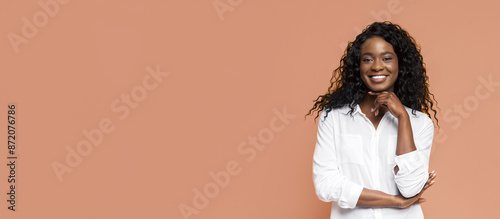 African American woman with long, curly black hair, wearing a white button-up shirt. She is smiling and has her arms crossed in front of her. The background is a peachy-orange color. photo