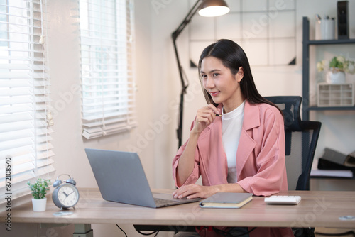 Woman working from home on a laptop, thoughtfully looking at the screen, in a cozy, well-lit room with office supplies on the desk.