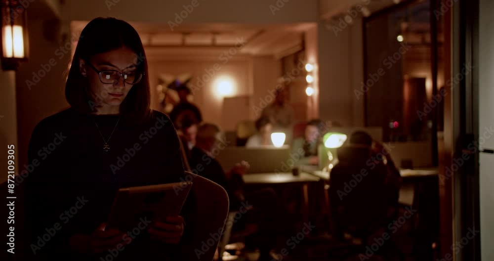 In a well-lit workspace after hours, a focused female employee works on a project, searching for information on her tablet, demonstrating her dedication and commitment to her work. In a well-lit workspace after hours, a focused female employee works on a project, searching for information on her tablet, demonstrating her dedication and commitment to her work.