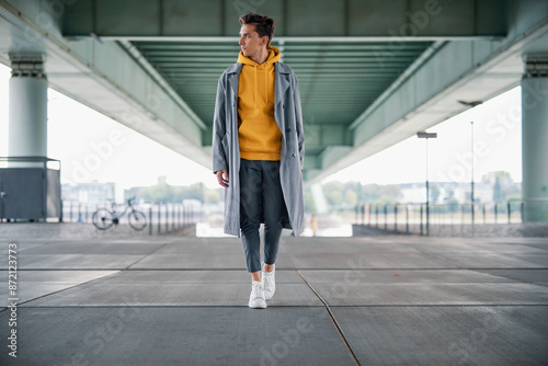 Young Man in Yellow Hoodie with Coat Walking Under Bridge on Sidewalk While Looking to the Left Side