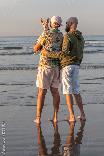 gay couple back to back with their dog on their lap looking at the camera, on the beach