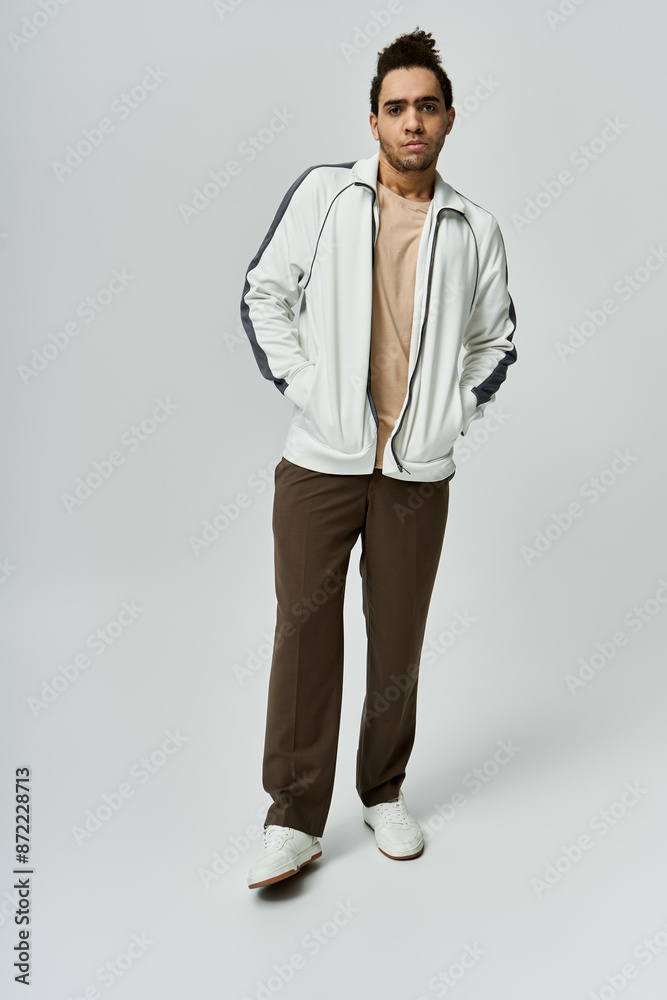 A young African American man wearing a white tracksuit with black accents poses against a grey backdrop. A young African American man wearing a white tracksuit with black accents poses against a grey backdrop.