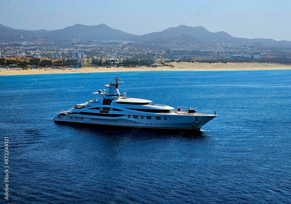 Obraz premium Luxury yacht sailing near the coastline with cityscape and mountains in the background.
