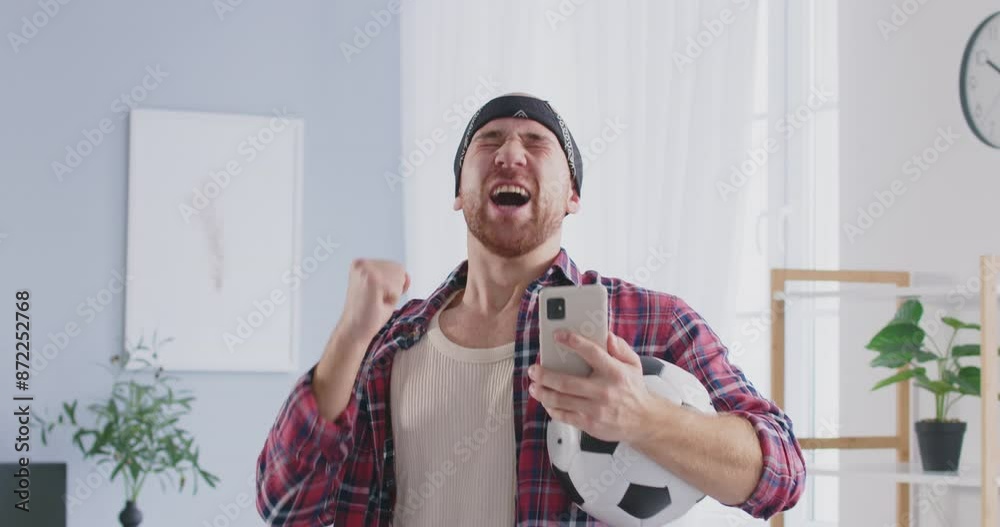 Happy young funny excited guy with soccer ball using smartphone, winning sports bet standing at home. Sports fan man watching football match on smartphone. 4k video. Slow motion video. Happy young funny excited guy with soccer ball using smartphone, winning sports bet standing at home. Sports fan man watching football match on smartphone. 4k video. Slow motion video.