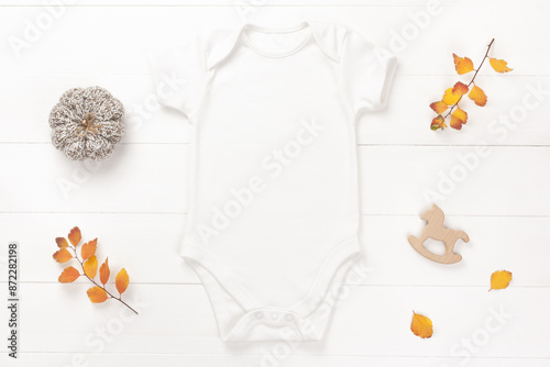 Autumn mockup of white baby bodysuit on wood background with yellow leaves, seasonal decor and toys, flat lay. Blank baby clothes template.