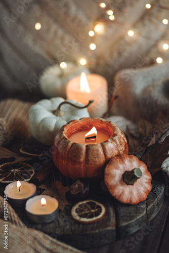 Cozy autumn home still life scene. Ceramic handmade pumpkin mug with burning candle near the window. Fall mood, Thanksgiving concept. Pumpkins, dry leaves, wool sweater, wooden background